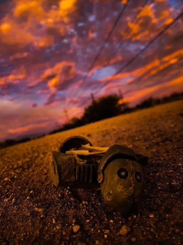 A small toy robot lying on its side in desert dirt at sunset, dramatic orange and purple clouds with power lines overhead.