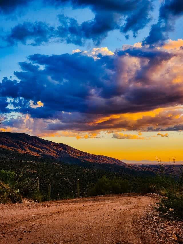A dirt road curves through a desert valley at sunset, mountains catching gold light, dramatic blue and amber clouds overhead.
