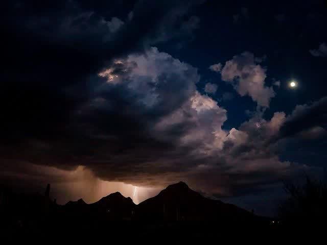 A lightning bolt strikes behind dark mountain silhouettes under churning night storm clouds, a bright moon glowing at upper right.