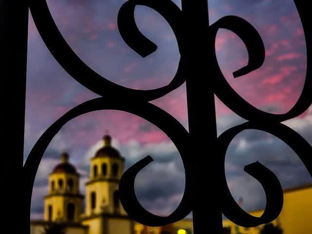 The twin bell towers of Mission San Xavier del Bac seen soft and golden through the dark curls of an iron gate, pink dusk clouds above.