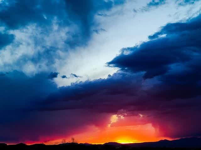 Huge blue and violet monsoon clouds stacked above a glowing orange horizon where the sun sets behind dark mountains.
