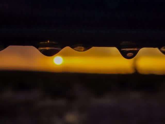 Close-up of water droplets clinging to a wire, the setting sun glowing gold behind them, a desert landscape softly blurred below.