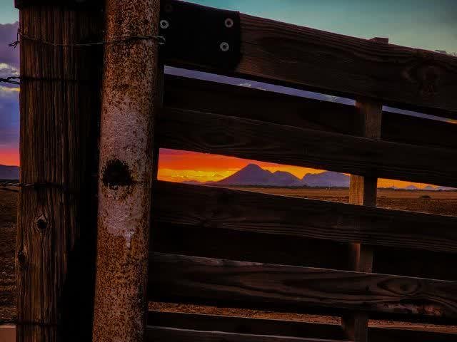 A weathered wooden ranch gate in silhouette framing a blazing orange sunset over a desert plain and distant mountain peaks.